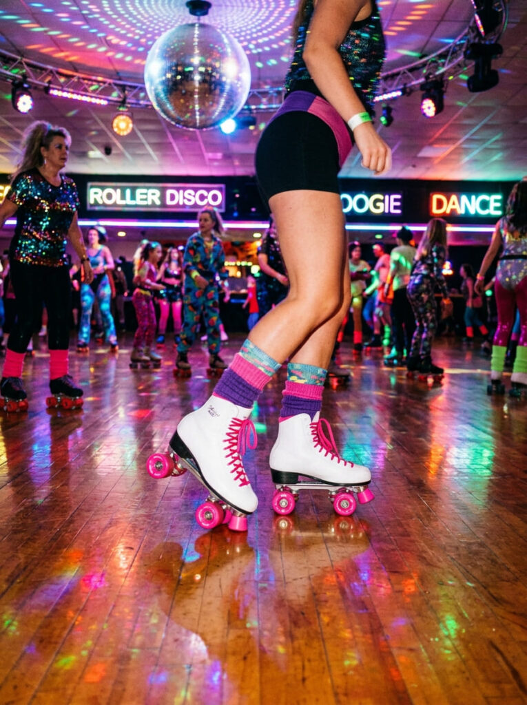 A close-up shot of white quad roller skates on a wooden rink floor with colorful retro disco lighting reflections.