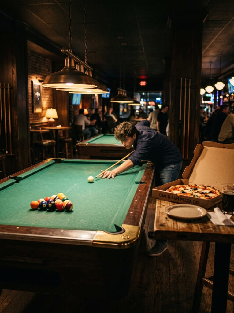 A 19-year-old guy lines up a pool shot under low-hanging lamps next to pizza boxes.