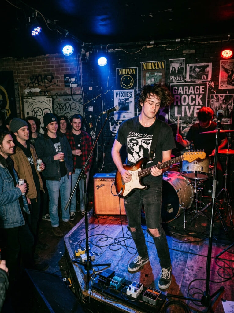 A 19-year-old guy plays electric guitar on a small stage under deep red and blue spotlights.